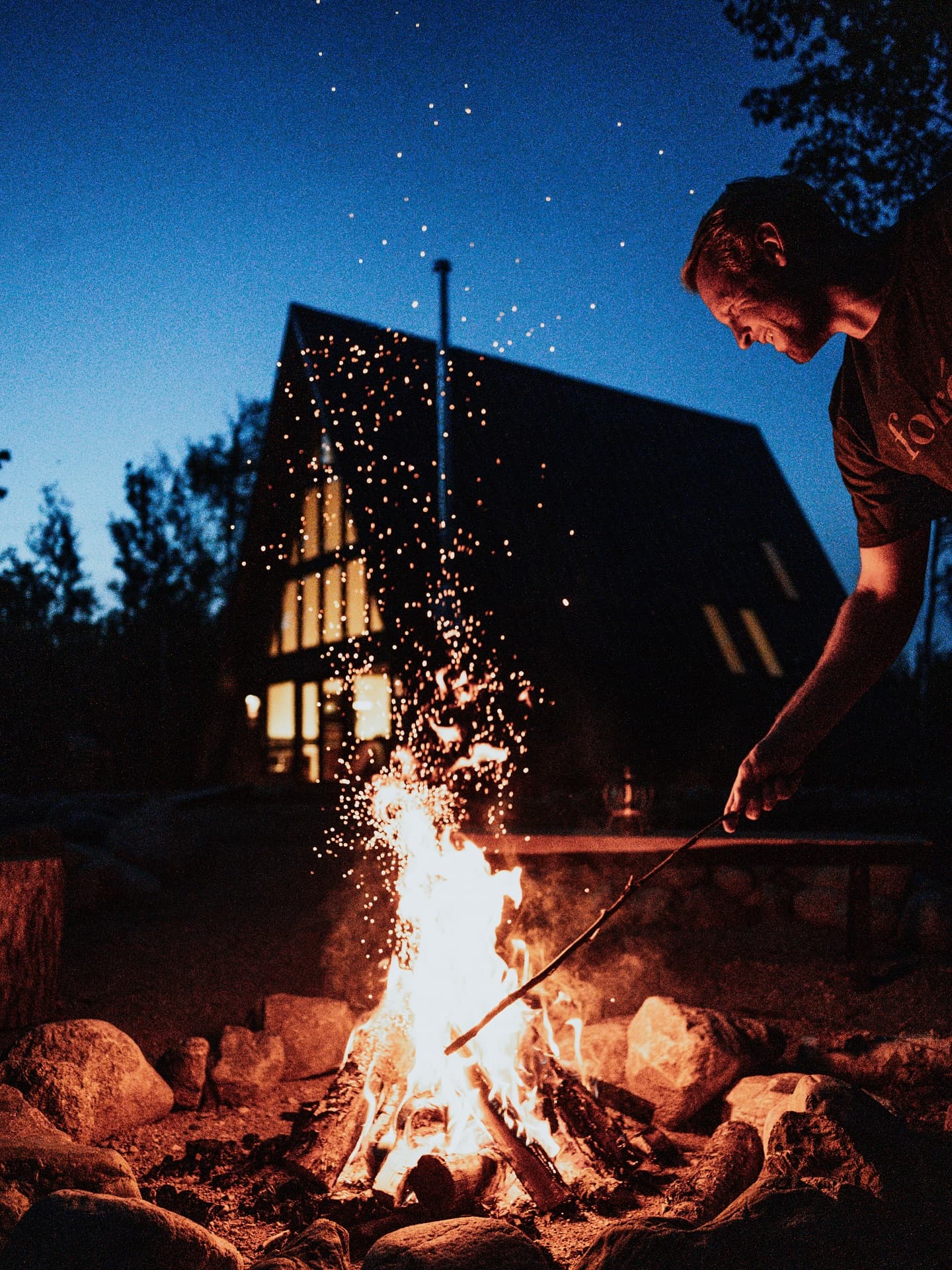 Person poking a campfire at night in front of an illuminated A-frame cabin