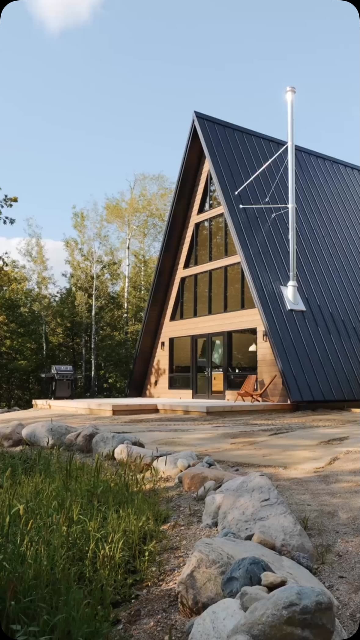 A-frame cabin with black metal roof surrounded by birch trees and boulders