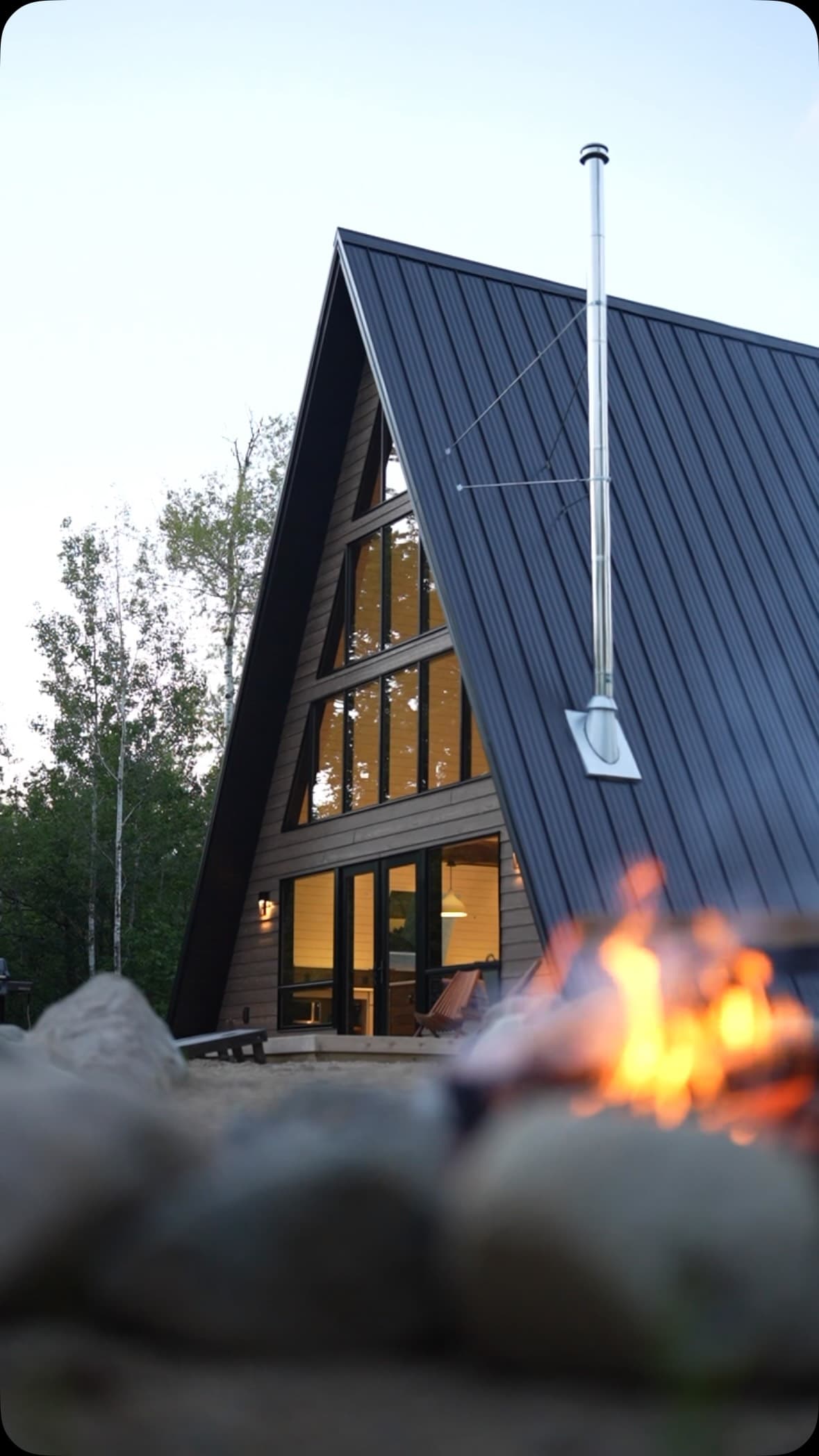 Modern A-frame cabin with glowing windows and firepit at dusk